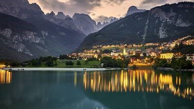 View of Trento valley at dust, with city lights reflected on the lake in the foreground, and mountains in the background